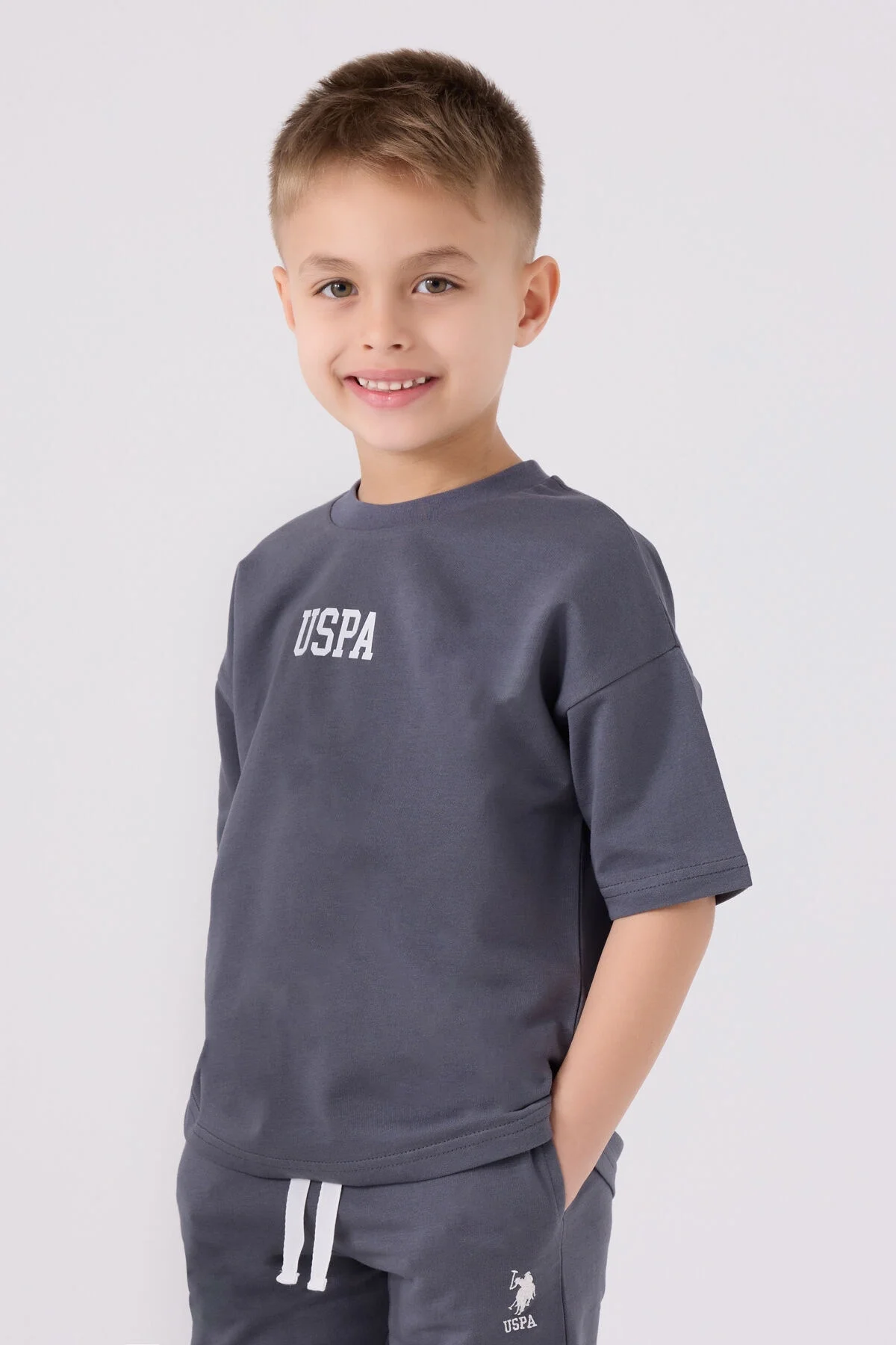 Antrasit renkli U.S. Polo Assn erkek çocuk Bermuda takım, ön ve yan görünüm, çocuk modeliyle gösterilmiştir.