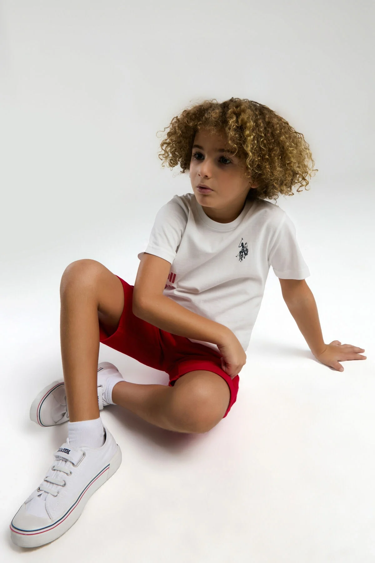 Beyaz U.S. Polo Assn erkek çocuk Bermuda takım elbette, kırmızı detaylı, marka logosu ve U.S.P.A. yazısı