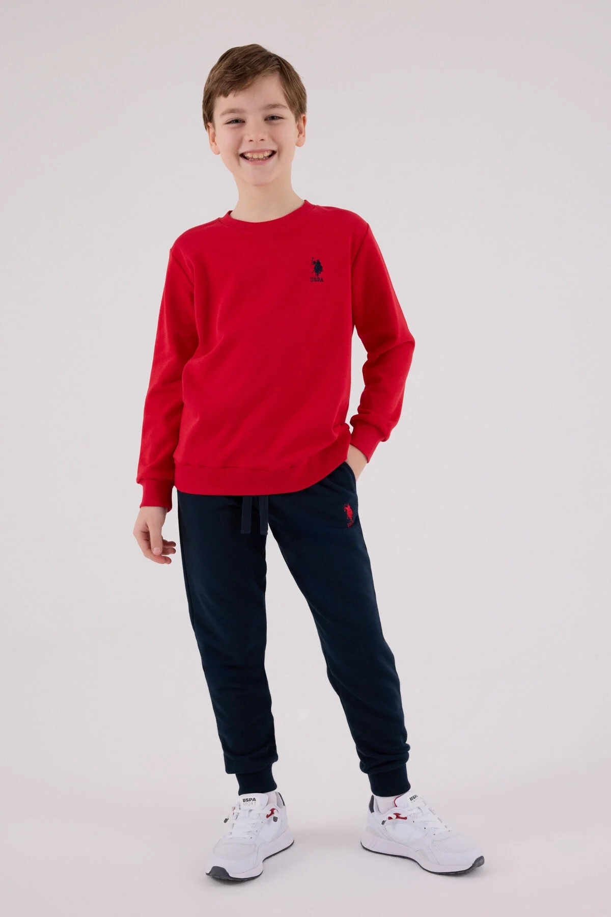 Kırmızı U.S. Polo Assn erkek çocuk eşofman takımı, model ön görünüm, siyah pantolon ve beyaz spor ayakkabıyla
