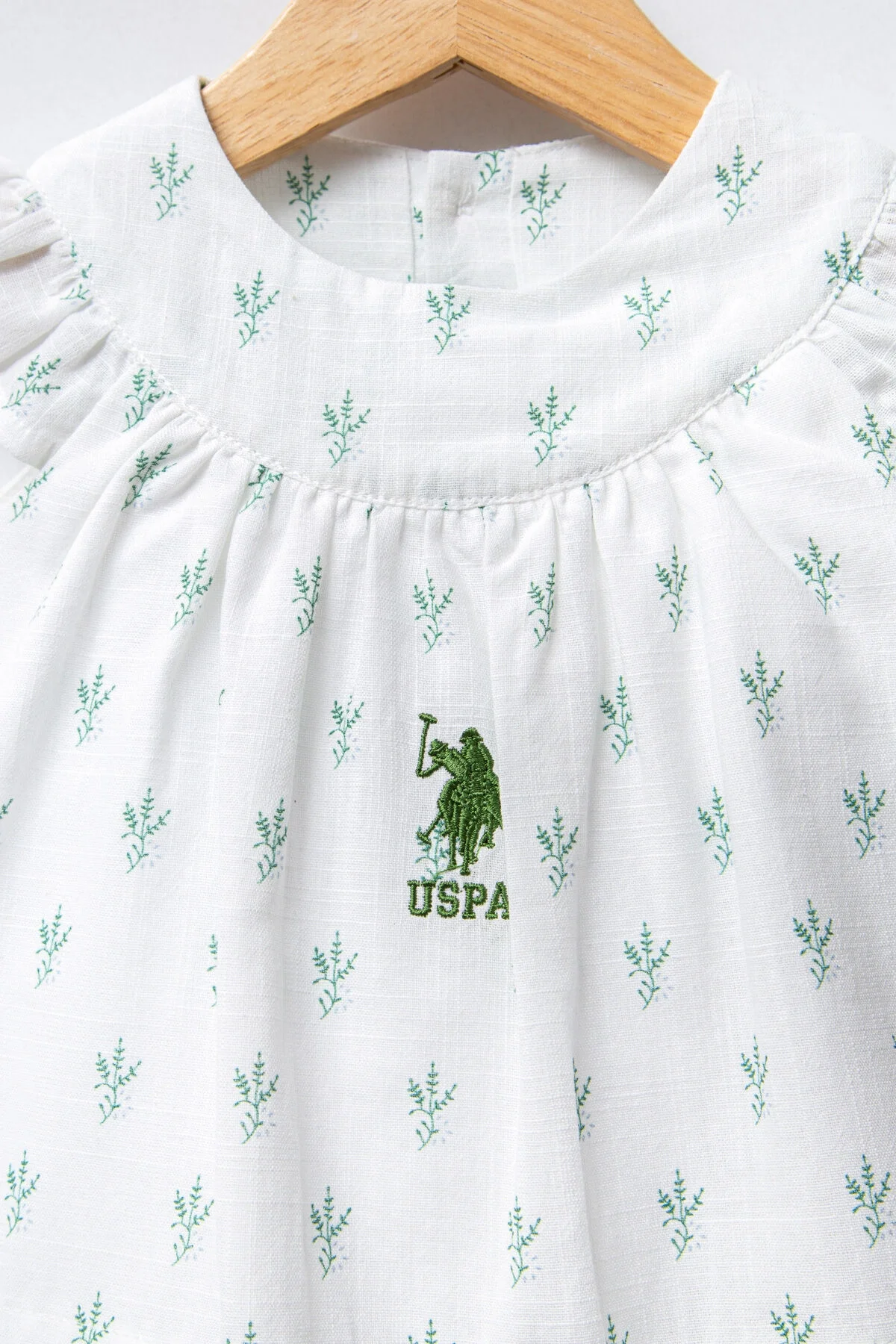 Beyaz desenli U.S. Polo Assn kız bebek elbise, kolları olmayan, göğüs detayı, marka logosu, arka plan: beyaz duvar