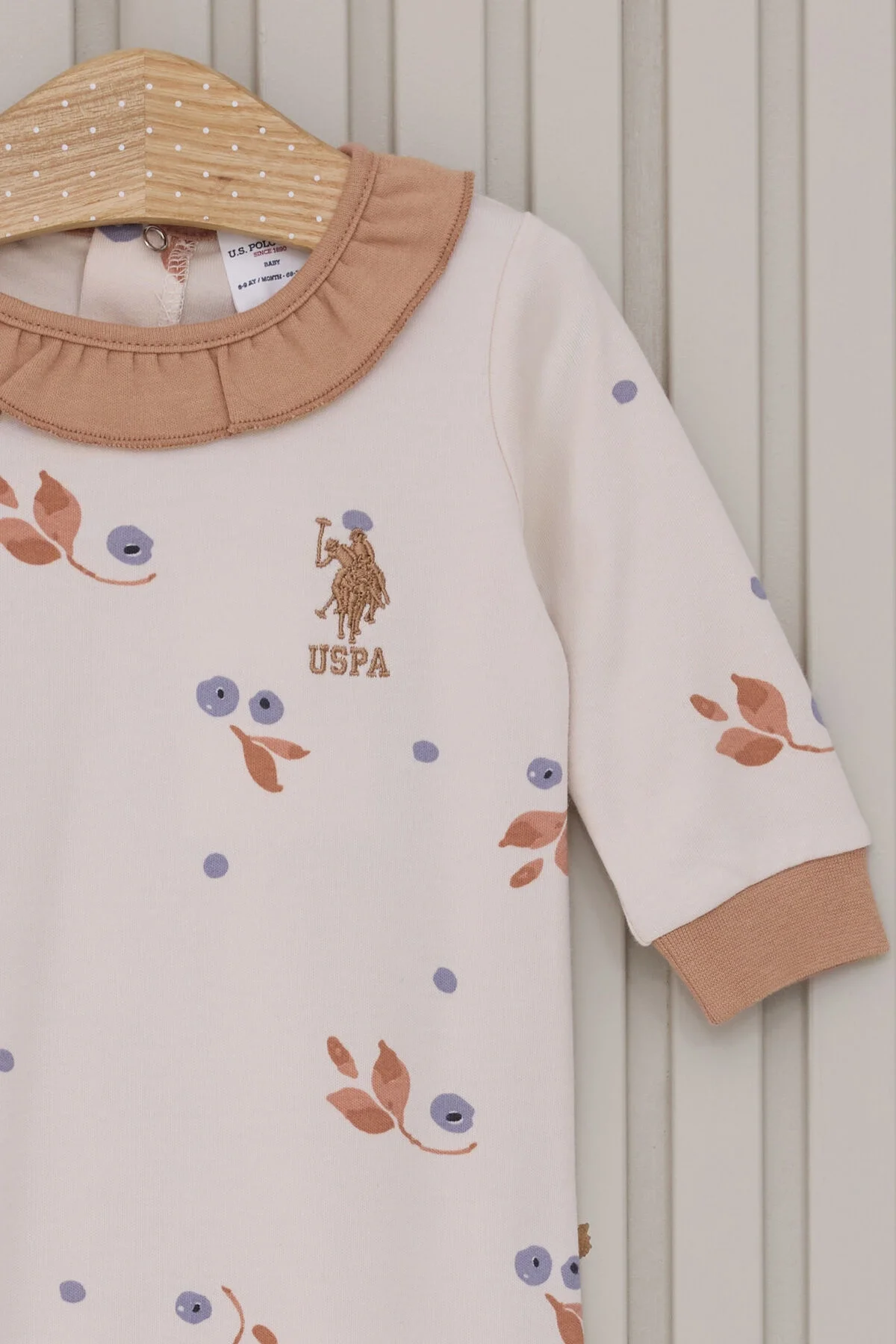 U.S. Polo Assn Kız Bebek Krem Tulum, krem zeminde mavi ve kahverengi yaprak deseni, kollar ve bacaklar kahverengi detaylı, ön görünüm