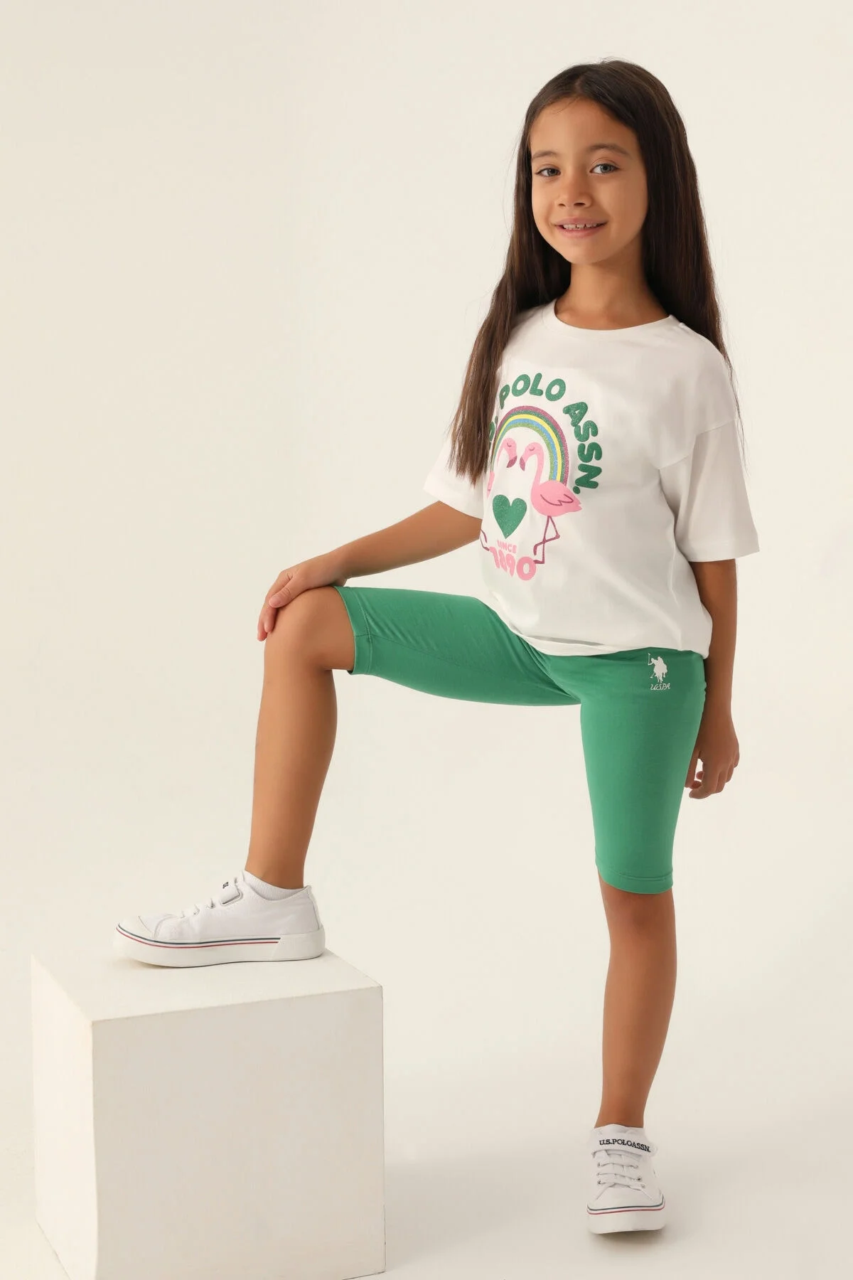 Beyaz flamingo ve rainbow desenli U.S. Polo Assn kız çocuk Bermuda takım, yeşil biker pantolon ve beyaz spor ayakkabıyla tamamlayıcı