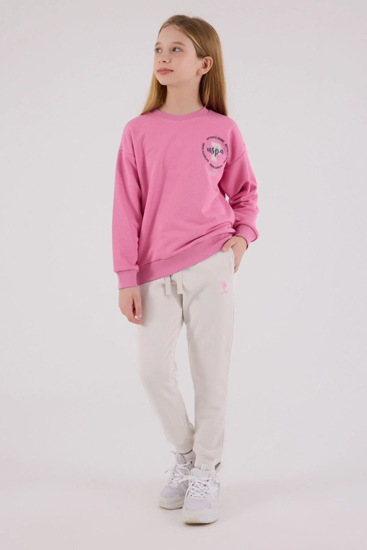 Pembe U.S. Polo Assn kız çocuk eşofman takımı, model ön görünüm, beyaz pantolon ve spor ayakkabı kombinasyonu
